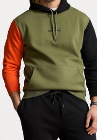 Mężczyzna w oliwearowej zielonej, czarnej i pomarańczowej bluzie z kapturem w kolorze blokowym, z kieszenią na przodzie oraz czarnych spodniach dresowych, ręce swobodnie opuszczone wzdłuż ciała.