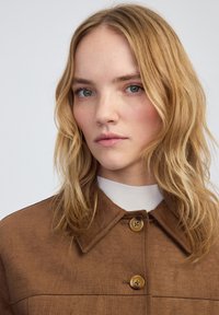 Chaqueta marrón texturizada con cuello, dos botones y una suave capa interior blanca. La tela presenta un sutil patrón de cocodrilo.