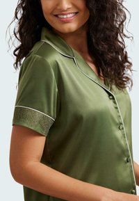 Top in satin verde oliva con chiusura a bottone, piping bianco e dettagli in pizzo sulle maniche corte. Tessuto liscio e vestibilità comoda.