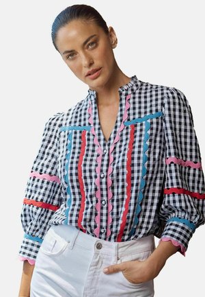 Femme aux cheveux tirés en arrière portant un chemisier à carreaux noir et blanc avec des bordures en galon rose, rouge et bleu, et un pantalon blanc, main dans la poche.