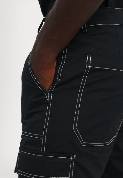 Black cargo pants with white stitching. Features pockets on the sides and a fitted design. Material appears durable with a smooth texture.