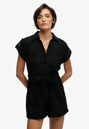 Vrouw met kort donker haar, gekleed in een zwarte, korte, met ceintuur omgeslagen romper met voorzakken, staand met handen achter haar rug.