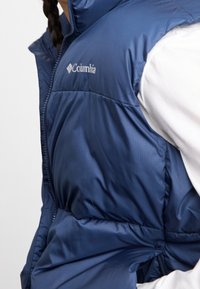 Tmavě modrá zateplená vesta z lesklého nylonu, která má vysoký límec, přední zip a logo Columbia na hrudi.