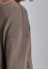 Långärmad plagg i mjukt, dämpat taupe-tyg. Har en rundad krage och avslappnad passform, med synlig sömdetalj på axeln.
