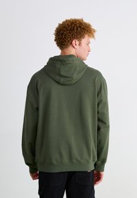 Sudadera verde oliva con capucha, de corte relajado, con puños y dobladillo de canalé. Textura suave con mínimos detalles, llevada al revés.