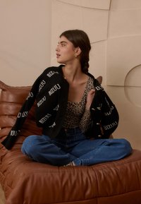 Jonge vrouw met gevlochten haar zit met gekruiste benen op een bruine leren bank, draagt een top met luipaardprint, blauwe spijkerbroek en een zwarte cardigan met witte tekst.