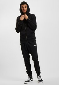 Felpa nera con cerniera e texture a coste, abbinata a pantaloni jogger coordinati con un logo bianco. Il modello indossa sneaker alte nere.