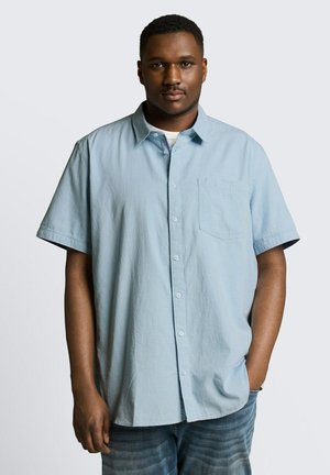 Man with short hair wearing a light blue short-sleeve button-up shirt and blue jeans, standing with one hand in pocket.