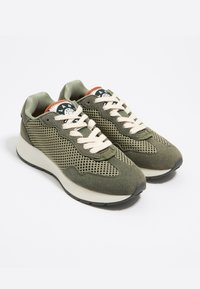 Sneaker verde oliva realizzati in materiali mesh e suede, con lacci crema e una suola testurizzata. Logo circolare distintivo sulla linguetta.
