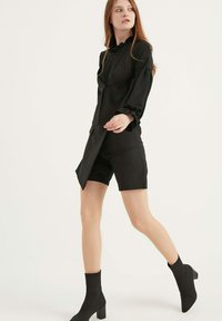 Zwarte jurk met gerimpelde kraag, lange mouwen en een gespleten voorkant, gecombineerd met zwarte strak zittende shorts en enkellaarsjes. Glad textuur.