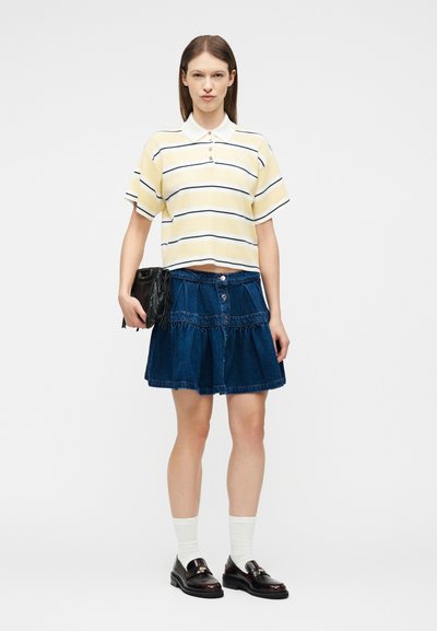 Νεαρή γυναίκα που φοράει πουκάμισο πόλο με κίτρινες και λευκές ρίγες, μπλε τζιν φούστα, λευκές κάλτσες, μαύρα μοκασίνια και κρατά μια μαύρη τσάντα.