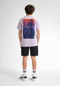 Lilla T-shirt med korte ærmer med et grafisk print på ryggen. Matchet med sorte shorts, hvid- og sort sneakers og stribede sokker.
