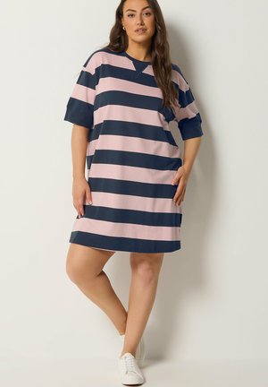 Donna che indossa un vestito corto a maniche corte, ampio, con larghe strisce orizzontali blu navy e rosa chiaro, in piedi davanti a uno sfondo bianco uniforme.