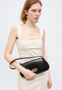 Νεαρή γυναίκα με αμάνικο κρεμ φόρεμα κρατώντας μια μαύρη δερμάτινη τσάντα με χρυσό κούμπωμα, μπροστά σε απλό ανοιχτό φόντο.