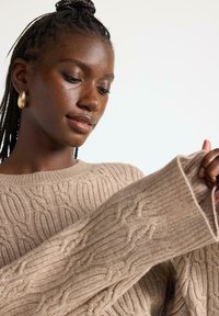 Maglione beige a maglia cable con polsini a coste e scollatura rotonda, caratterizzato da motivi testurizzati e un aspetto morbido e accogliente.