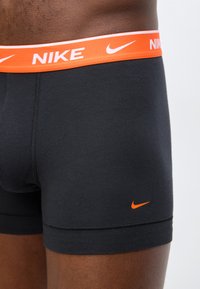 Gros plan sur une personne portant un boxer Nike noir avec une ceinture orange et un logo swoosh Nike orange sur la cuisse gauche.