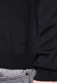 Maglione nero lavorato a maglia con un motivo testurizzato, polsini a costine e una piccola etichetta del marchio sul lato inferiore. Abbinato a jeans grigi.