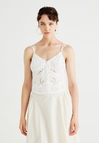 Vit bomullscamisole med justerbara spaghetti-remmar, med broderade mönster och knäppning fram. Slät textur, delikat design.