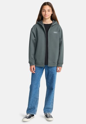 Un adolescent portant un sweat à capuche zippé vert foncé, un t-shirt noir, un jean bleu et des baskets noires avec des semelles blanches, se tenant devant un fond uni.