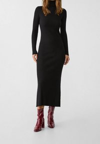 Robe noire en tricot côtelé avec des manches longues et un col roulé, présentant une coupe slim et un ourlet droit, assortie à des bottines bordeaux brillantes.
