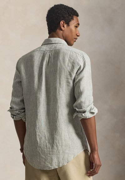 Férfi feltűrt ujjú, világosszürke és fehér függőleges csíkos ingben, bézs nadrágban, semleges háttér előtt áll.