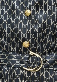 Tecido azul-marinho com padrão repetido de logotipos brancos. Acessórios de botão dourado e uma fivela circular dourada num detalhe do cinto. Textura lisa.