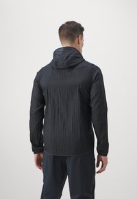 Veste légère noire avec une finition texturée, capuche à cordon de serrage et manches longues, vue de dos. Design simple, sans logos visibles.