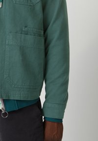 Grüne Canvasjacke mit leicht übergroßer Passform, ausgestattet mit einer Brusttasche und gerippten Bündchen, über ein blaues Reißverschluss-Basisschicht getragen.