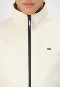 Light beige zip-up jacket with high collar, black zipper, and small embroidered logo featuring blue, white, and red accents on the chest.