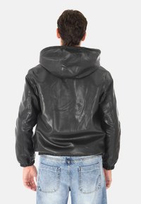 Uomo che indossa una giacca di pelle nera con cappuccio e jeans in denim azzurri, di spalle su uno sfondo semplice.