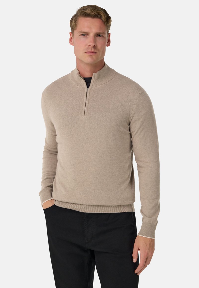 Pull à fermeture éclair beige en matière douce, doté d'un col montant et de poignets côtelés, associé à un pantalon noir pour un look superposé.