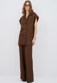 Blazer senza maniche marrone doppiopetto con grandi bottoni, abbinato a pantaloni larghi marroni. Entrambi realizzati in tessuto liscio, con tasche laterali.