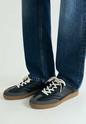 Zapatillas negras y grises con cordones blancos, con suela de goma texturizada y diseño elegante. Combinadas con vaqueros de mezclilla azul oscuro.