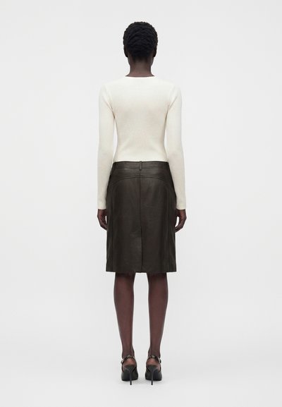 Femme debout de dos, portant un haut blanc à manches longues, une jupe en cuir sombre jusqu'aux genoux et des chaussures à talons noirs.