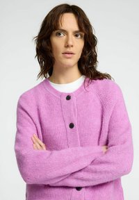 Rosa gestrickte Strickjacke mit einer weichen Textur, runder Halslinie und zwei schwarzen Knöpfen vorne. Arme vor der Brust verschränkt.