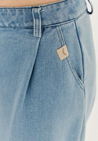 Svetlomodré denimové nohavice majú jednoduchý dizajn, s viditeľnou kapsou a malou béžovou nášivkou s líniovým detailom.