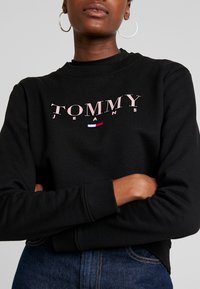 Černá bavlněná mikina s logem "TOMMY JEANS" v růžové barvě, s malým modro-bílým akcentem pod logem. Žebrované man�žety a lem.