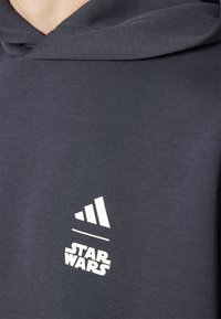 Temno siva kapuca s kapuco, izdelana iz mehkega blaga. Ima bele natisnjene logotipe: tri črte in 'STAR WARS' blizu roba.