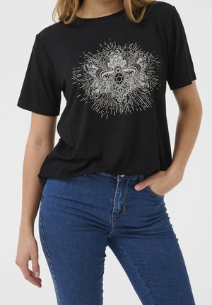 Kobieta nosząca czarną koszulkę z misternym białym kwiatowym i geometrycznym wzorem mandali, w połączeniu z niebieskimi dżinsami, ręka w kieszeni.