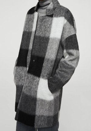 Manteau à carreaux en mélange de laine noir, blanc et gris, manches longues, col et poches latérales, porté sur un pull à col roulé gris.