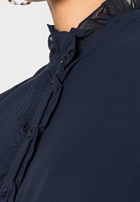 Námořnická modrá blůza s volánkovým límcem, svislými záhyby a černými knoflíky vpředu. Hladká textilie, detailní pohled.