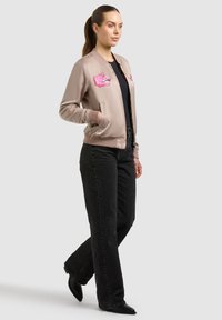 Femme aux cheveux bruns attachés en queue de cheval, portant une veste bomber beige avec des patchs tigre roses, un haut noir, un jean large noir et des chaussures noires pointues.