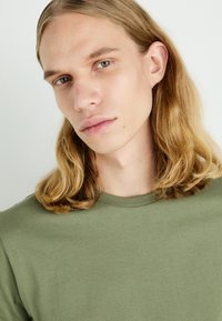 Miękka zielona bawełniana koszulka z okrągłym dekoltem. Modelka ma długie, falujące włosy i jasną skórę, częściowo widoczna w ujęciu zbliżeniowym.