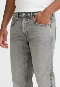 Jean en denim gris avec une finition délavée, conception standard à cinq poches et une fermeture à bouton métallique à la ceinture. Tissu texturé visible.