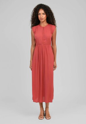 Femme aux cheveux bouclés portant une robe midi corail sans manches et des talons nus à brides, debout devant un fond gris clair uni.