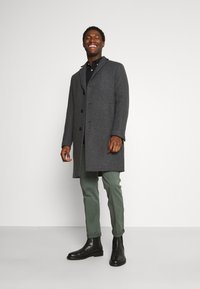 Gray, wool-blend overcoat with a collar, front buttons, and side pockets, worn over a black shirt, paired with green pants and black boots.
