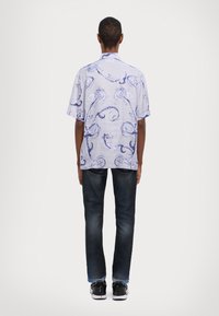 Camisa de manga corta en azul claro con patrones oscuros en espiral, combinada con jeans oscuros y zapatillas negras. Vista desde la parte de atrás.