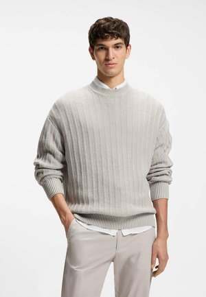 Giovane uomo con capelli corti e scuri, indossa un maglione grigio chiaro a coste sopra una camicia bianca e pantaloni beige, in piedi con una mano in tasca.