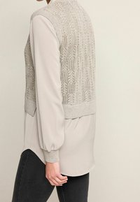 Gilet a maglia kaki con retro decorativo a fantasia, abbinato a una blusa crema a maniche lunghe. Tessuto morbido, vestibilità comoda e polsini a coste.