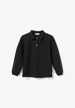 Uśmiechnięty młody chłopiec z blond włosami, ubrany w czarne, długorękawowe polo, niebieskie dżinsy i białe trampki, stojący z lekko uniesionymi ramionami.